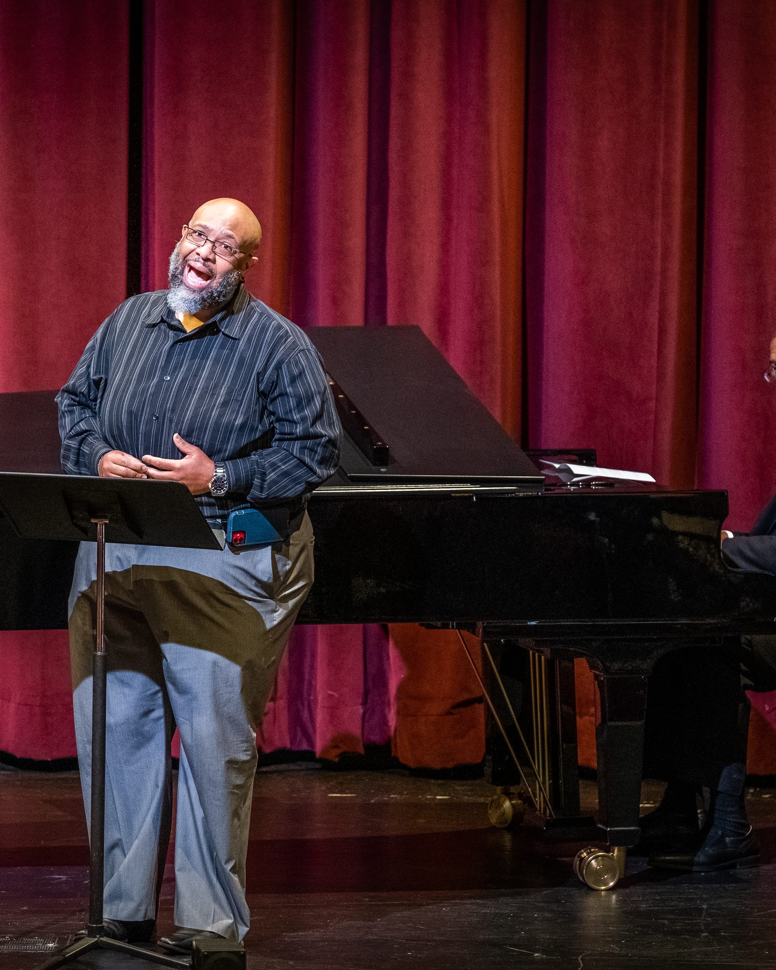 Singer gesturing on stage while a pianist performs against red theatre curtains