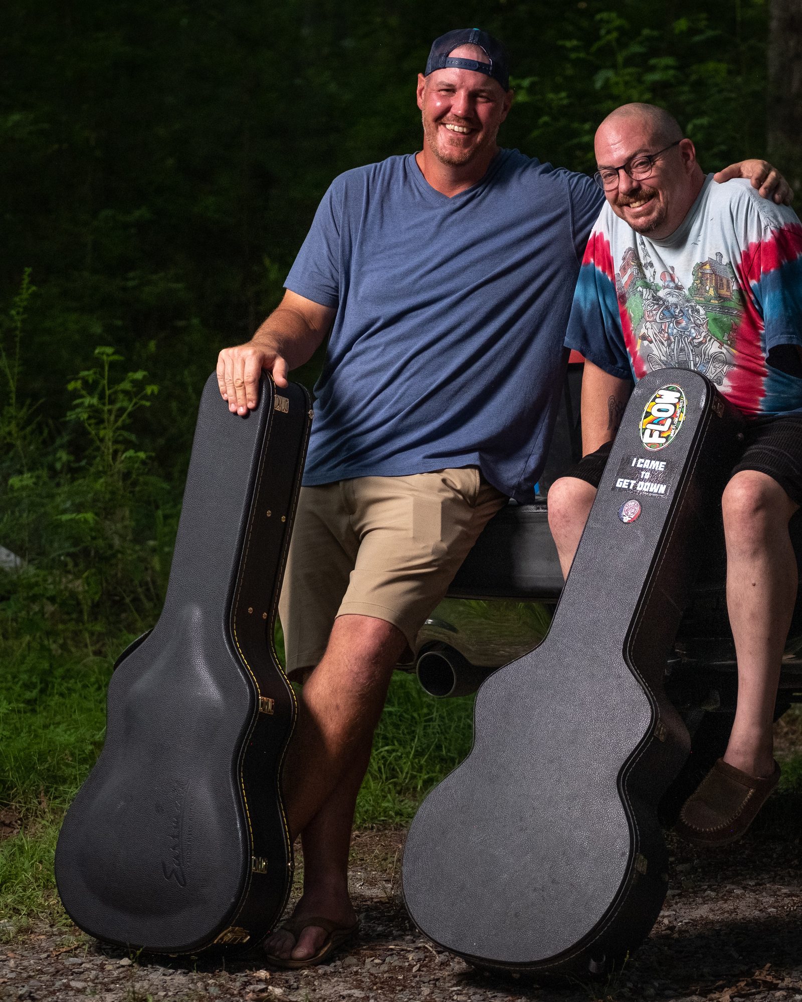 Two musicians posing outdoors beside a pickup truck with their instrument cases