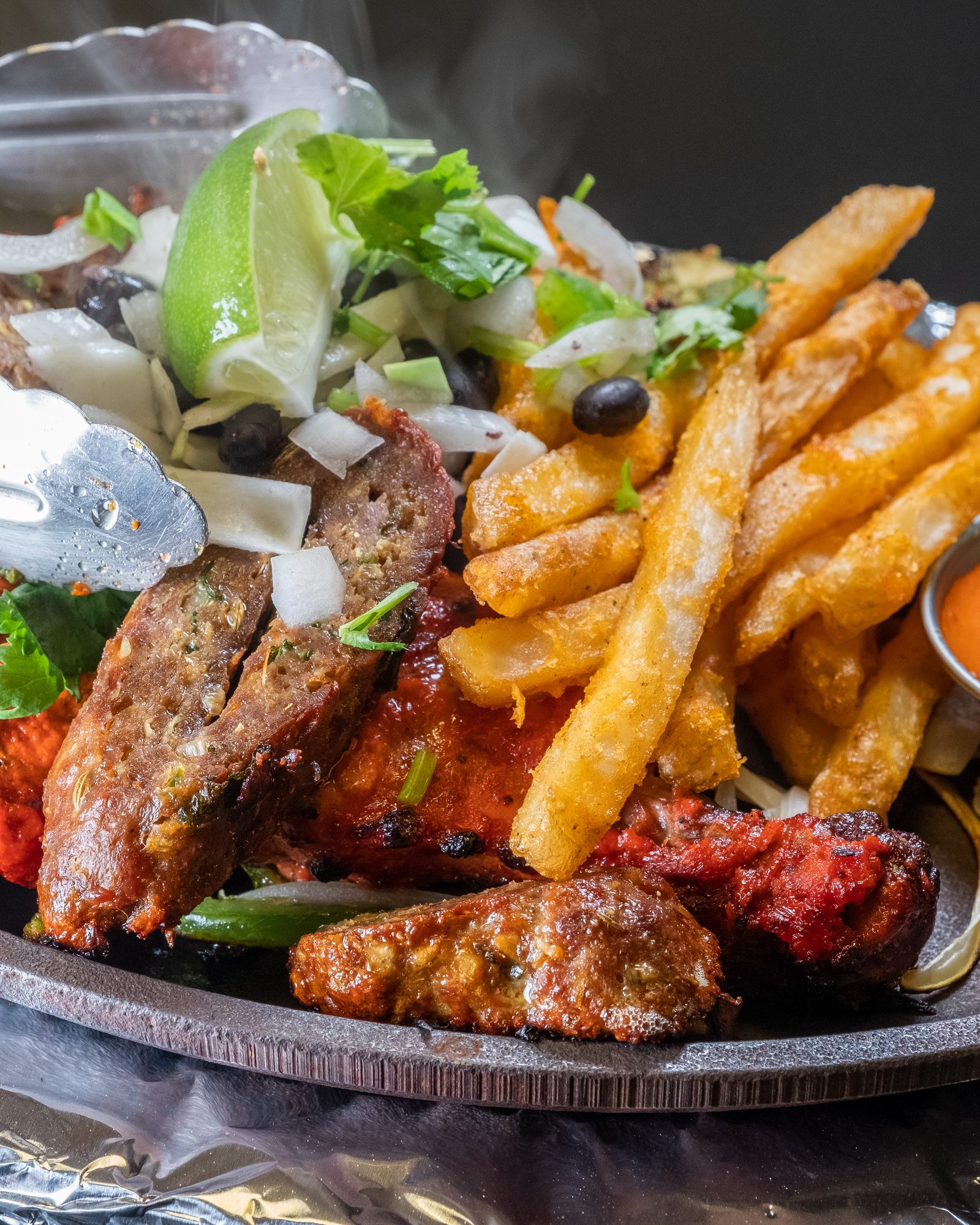 Steaming mixed grill platter with fries, vegetables, lime, and sauces